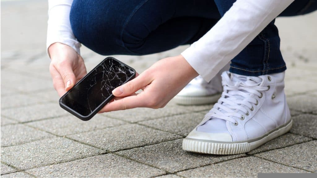 imagem mostra uma pessoa recolhendo do chão um celular com a tela quebrada